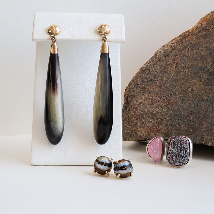 Black and white drop earrings with gold tops hanging on a white earring stand. Laying on the ground to the right of the earrings lies a pair of striped stud earrings and a pink and purple ring.