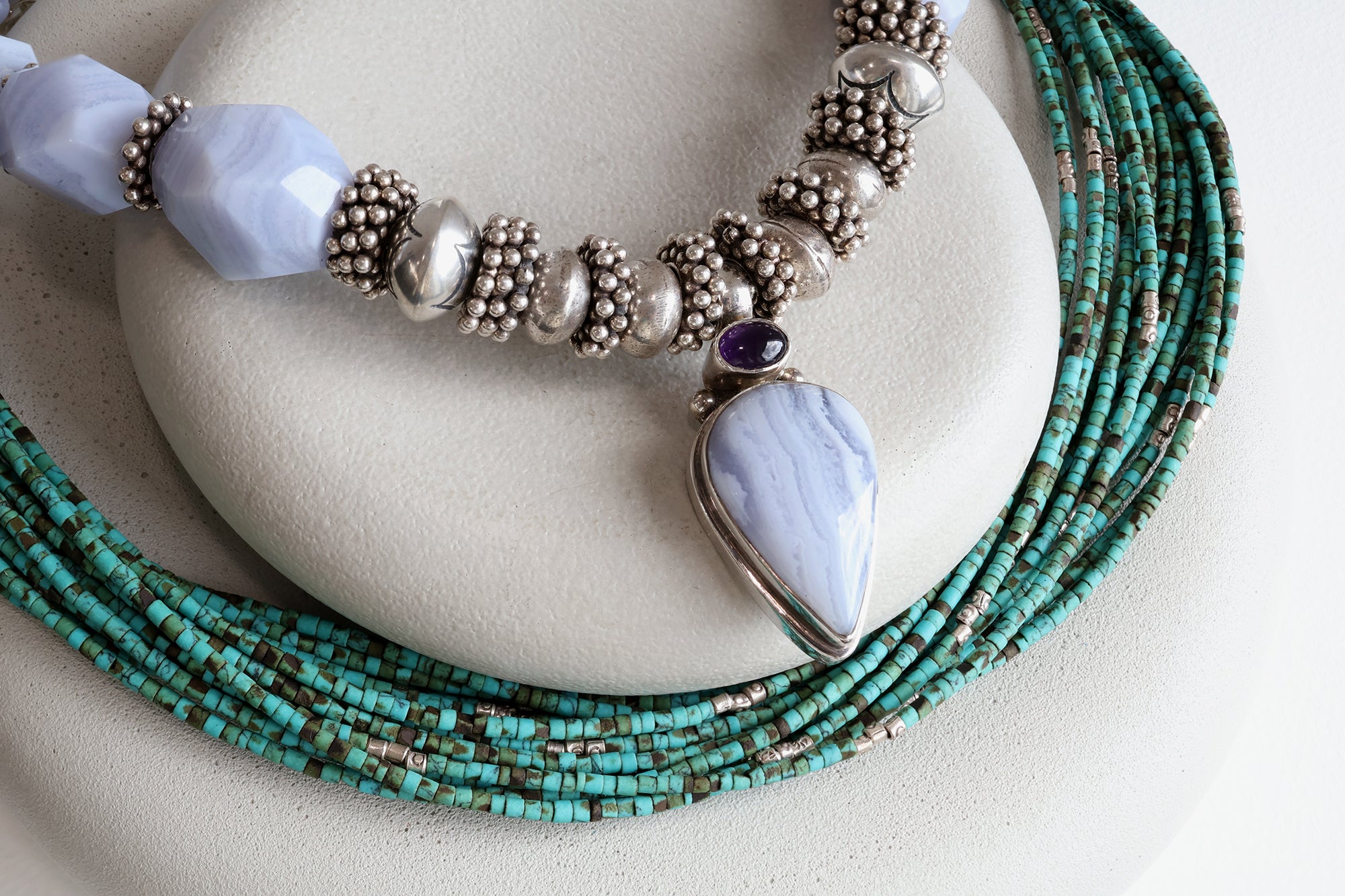Two necklaces on two levels of gray stones. The top necklace is a bold mixture of light blue stones and silver beads. The bottom necklace is a multi-strand necklace with dainty teal green beads.