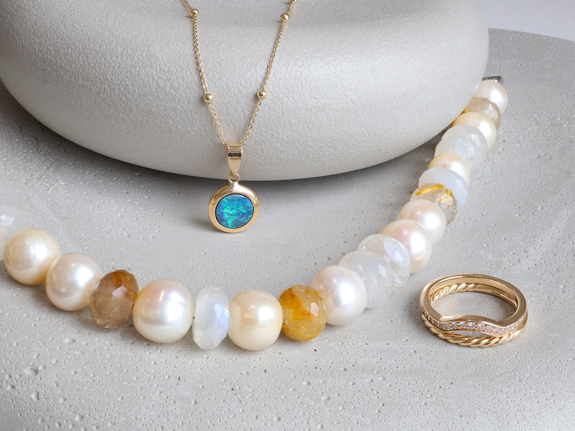 Opal necklace, pearl and gemstone beaded necklace, and gold ring set on grey stones.