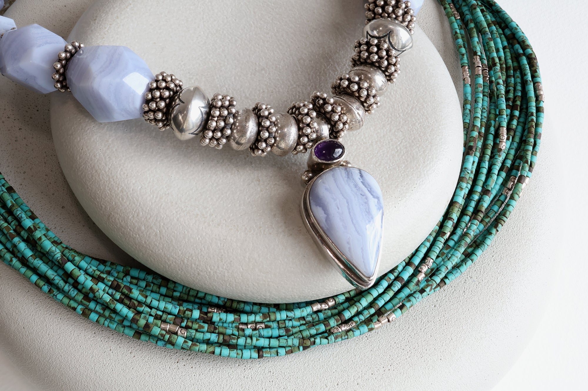 Two necklaces on two levels of gray stones. The top necklace is a bold mixture of light blue stones and silver beads.  The bottom necklace is a multi-strand necklace with dainty teal green beads. 