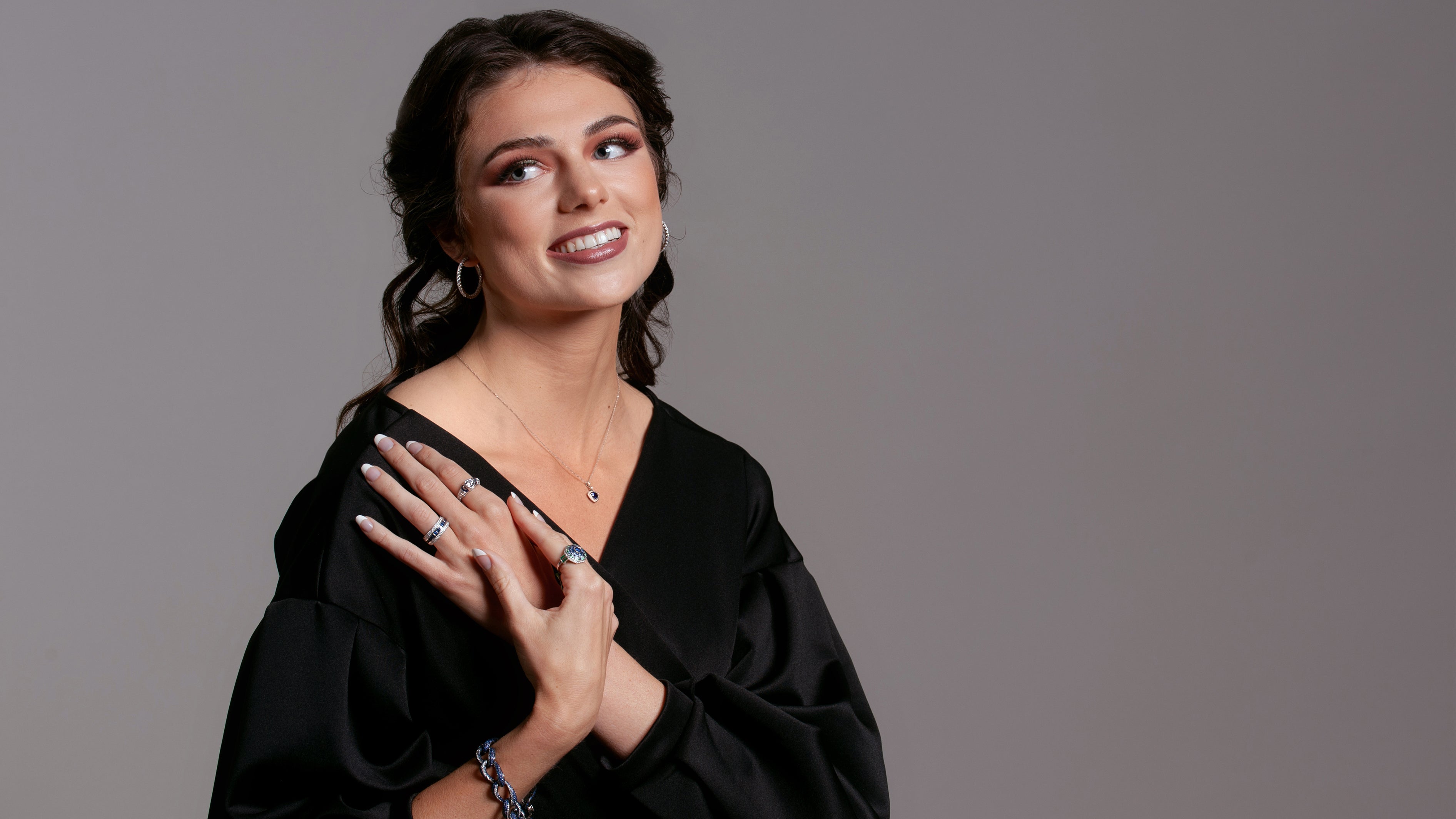 Brunette woman wearing black looking to the left.  Her hands are crossed and she is wearing blue gem rings, bracelet, and necklace with a pair of diamond hoops.