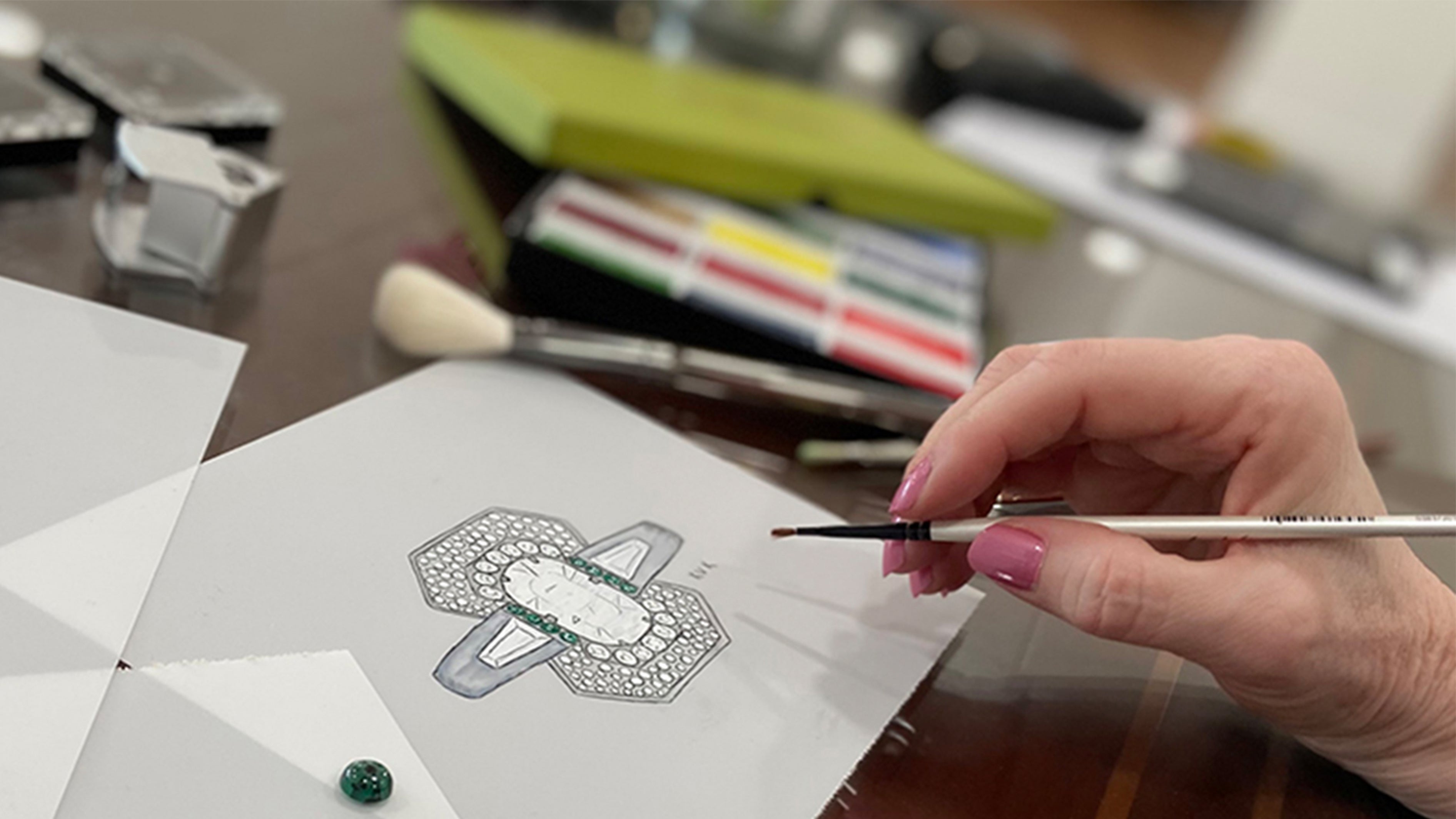 A hand holding a paint brush is finishing a painting of a custom design ring with a big oval diamond in the center and two tapered baguette diamonds on either side.  The desk has blurred paint brushes and paint colors in the background.