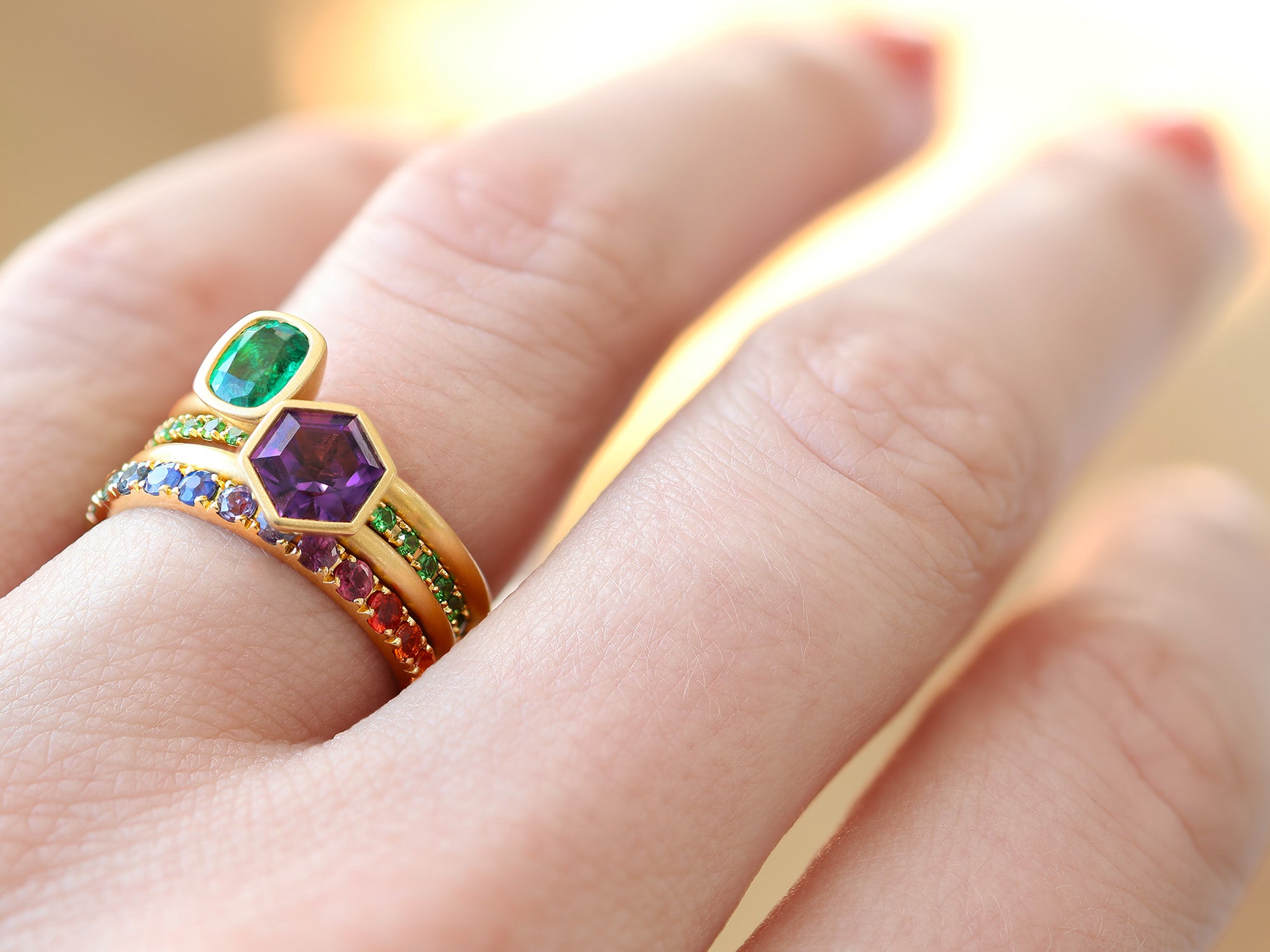 Oval emerald, hexagonal amethyst, and rainbow golden band stacked on a woman's ring finger.