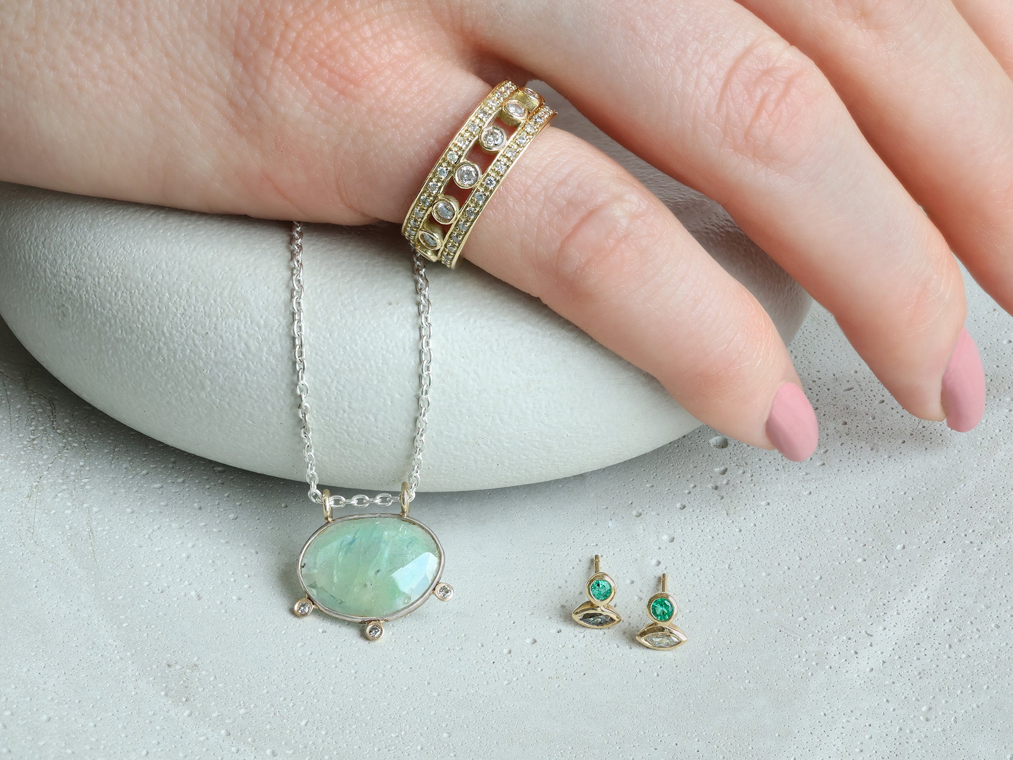 Women laying her hand on stones.  She's wearing a gold and diamond pinky ring while a necklace and two stud earrings are displayed below her hand.